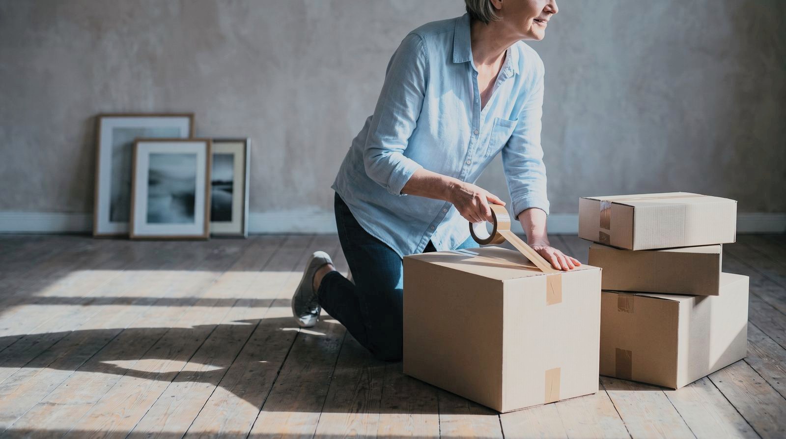 A woman taping a moving box shut in a half-empty living room